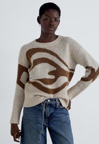 Beige strikkegenser med abstrakt brunt mønster, rund halsutskjæring, ribbestrikkede mansjetter og kant. Kombinert med blå jeans, uformet design.