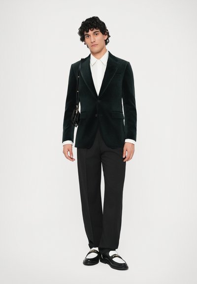 Sametový blazer tmavě zelené barvy s klopovými límci, zakončený bílou košili a černými kalhotami. Obsahuje malou černou crossbody tašku a boty.