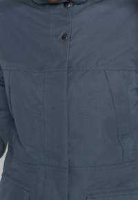 Veste bleu foncé, résistante à l'eau, dotée d'un col montant, d'une fermeture à boutons-pression et de deux poches avant avec coutures apparentes.