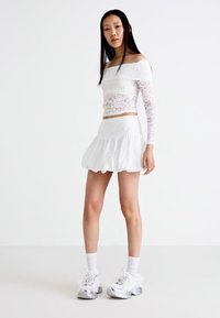 Crop top bianco in pizzo, senza spalle, abbinato a una mini gonna bianca con volants. La modella indossa sneakers argento chunky con calzini bianchi.