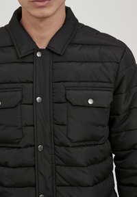 Sort polstret jakke med krave, snapknapper, to brystlommer og vandret quiltet struktur. Fremstillet af et let materiale.