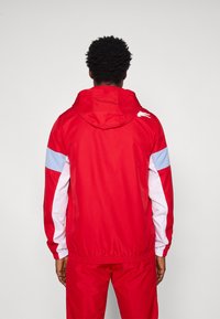 Κόκκινο τζάκετ με κουκούλα, με λευκές και μπλε λεπτομέρειες, ελαστική μέση και μαλακό, αδιάβροχο ύφασμα. Διαθέτει λογότυπο στην πλάτη.