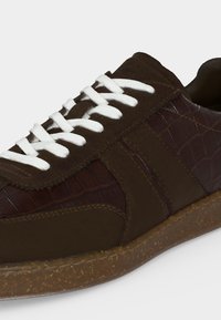 Baskets marron foncé avec lacets blancs, panneau latéral texturé ressemblant à une peau de crocodile, et semelle en gomme tachetée.