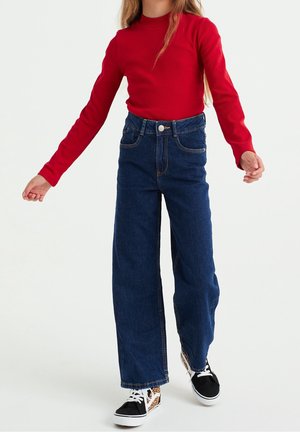 Dziecko ubrane w czerwoną bluzkę z długim rękawem, niebieskie szerokie spodnie z wysokim stanem oraz czarne trampki z panterkowymi detalami.