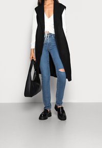 Chaleco negro sin mangas sobre una camiseta de rayas blanca, combinado con jeans ajustados azules con una rodilla desgastada. Mocasines de plataforma negros y bolso.