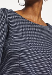 Marinblå stickad topp med rund halsringning, strukturerat mönster och framhävda axlar, med synlig sömnad längs kanterna.