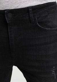 Pantalones vaqueros de denim negro con un diseño clásico de cinco bolsillos, un desvanecimiento sutil y un parche desgastado visible cerca de la rodilla.
