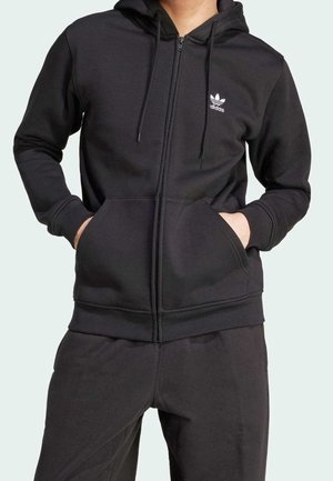 Person trägt schwarzen Adidas Kapuzenpullover mit Reißverschluss und passende schwarze Hose, Hände in den Taschen des Sweatshirts.