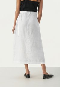 White linen midi skirt with a smooth texture, a belt at the waist, and a back slit. Worn with black flat shoes.