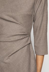 Robe en tissu à petits carreaux marron et beige, manches trois-quarts, avec un détail froncé et torsadé à la taille.