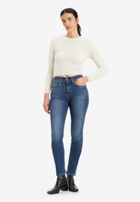 Pullover bianco corto con una finitura testurizzata abbinato a jeans blu scuro a vestibilità slim. Il modello indossa stivali neri fino alla caviglia. Design e vestibilità semplici.