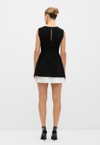 Robe noire sans manches avec une jupe plissée blanche et un détail de dos à découpe en forme de clé, associée à des chaussures à talons noirs. Texture de tissu lisse.