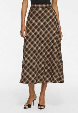 Femme portant une jupe midi trapèze à carreaux marron et beige, associée à des sandales noires à talons ouverts, debout sur un fond uni.