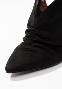 Stivale alto in suede nero con tacco a spillo e punta affusolata, caratterizzato da dettagli arricciati sulla parte anteriore, che mettono in mostra una texture liscia e una silhouette elegante.