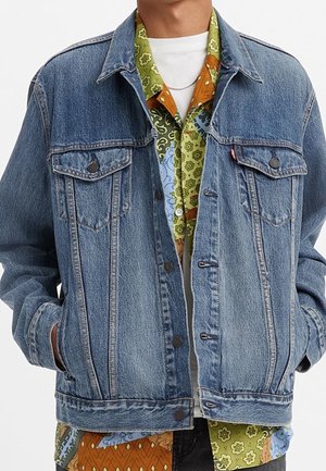 Person wearing light blue denim jacket over a multicolored patterned shirt and white t-shirt, hands in jacket pockets.
