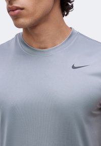 Homme portant un t-shirt Nike gris clair à col rond avec un logo swoosh noir sur la poitrine droite, montré du cou au haut du torse.