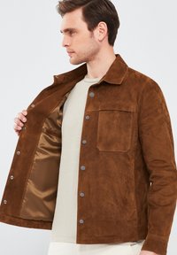 Bruine suede jas met een kraag in overhemdstijl, drukknoopsluitingen, één borstzak en een zijden beige voering. Subtiele textuur, getailleerde pasvorm.