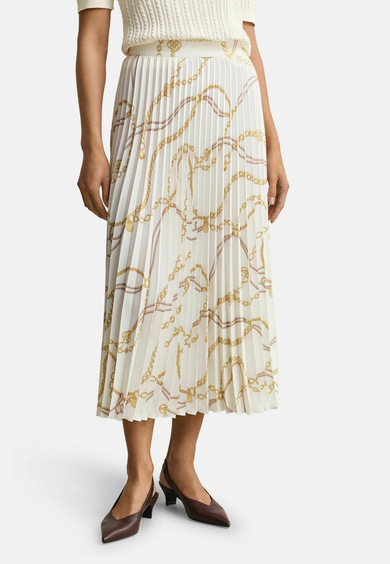 Femme portant une jupe midi plissée blanche avec des motifs de chaînes dorées et des chaussures slingback marron à petit talon, debout sur un fond uni.