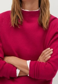 Fuchsia gestrickter Pullover mit hohem Kragen und gerippten Bündchen, der eine strukturierte Oberfläche hat und darunter ein mehrlagiges weißes Rüschenshirt trägt.