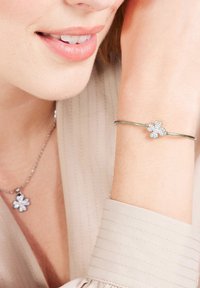 Zilveren armband met een bloemenvormige bedel versierd met heldere edelstenen; gedragen om een pols naast een soortgelijke bloemenhalsketting.