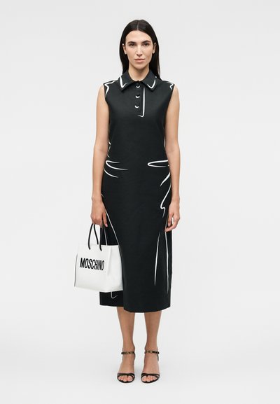 Γυναίκα με μαύρο μίντι φόρεμα χωρίς μανίκια με λευκές λεπτομέρειες περιγράμματος, κρατώντας λευκή τσάντα Moschino και φοράει μαύρες ψηλοτάκουνες με λουράκι στον αστράγαλο.