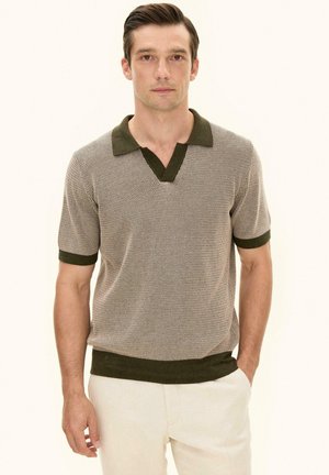 Mand iført beige kortærmet strikket polo med mørkegrøn krave, manchetter og kant, kombineret med lyse bukser, stående mod hvid baggrund.