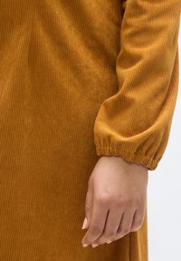 Ribb paradedress i rustfarge med kroppsnær overdel og voluminøse ermer, med elastiske mansjetter. Hånd hviler forsiktig mot stoffet.