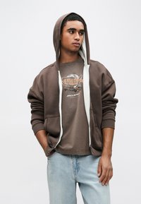 Hnedá mikina na zips so sivou podšívkou, doplnená o tričko s grafikou a svetlomodré džínsy. Voľný strih, mäkká textúra.