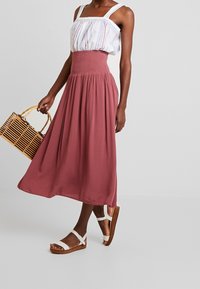 Zweiteiliges Outfit mit einem weißen, gestreiften Oberteil mit breiten Trägern und einem burgunderfarbenen, gerafften Maxirock. Die Accessoires umfassen eine Handtasche aus Bambus und weiße Sandalen.