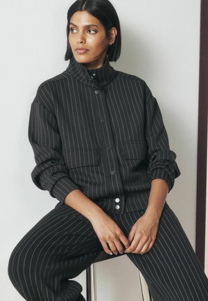 Woman with short dark hair wearing a black pinstripe jacket and pants, sitting on a stool, looking to the side with hands resting on her lap.