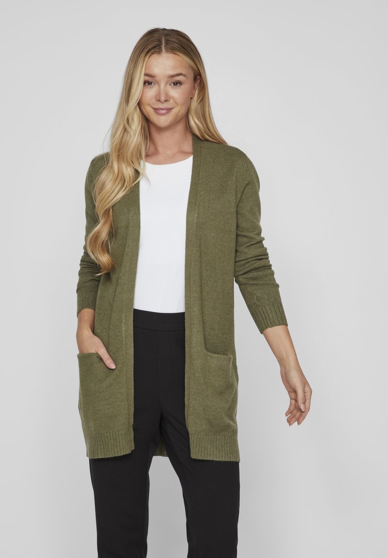 Long olive green cardigan made of soft, textured fabric. Features open front, ribbed cuffs, and two front pockets. Paired with a white top and black pants.