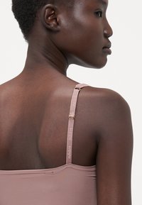 Light pink sports top with thin straps featuring adjustable hardware and branded text. Smooth, stretchy fabric with a seamless design.