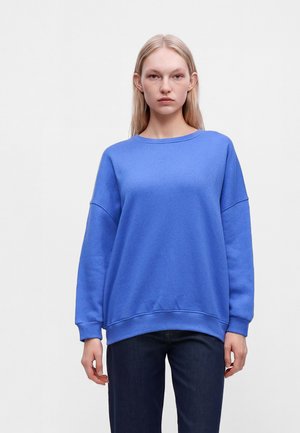 Vrouw met licht haar draagt een losse blauwe trui en donkere broek, staand tegen een effen lichtgrijze achtergrond.