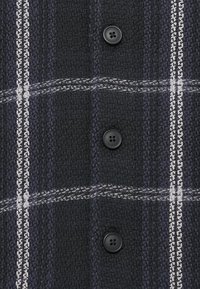 Tela texturizada en negro y azul oscuro con líneas de cuadro en blanco y gris, que presenta tres botones negros de cuatro agujeros dispuestos en fila vertical.