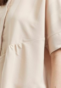 Světle béžová blůza s měkkou texturou, s nařasenými detaily, krátkými rukávy a zapínáním na knoflíky u výstřihu, se subtelnými vertikálními pruhy.