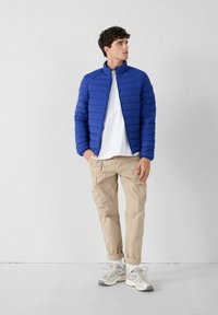 Man die staat met een blauwe gewatteerde jas, een wit T-shirt, beige cargobroek, witte sokken en grijs-witte sneakers tegen een plain muur.