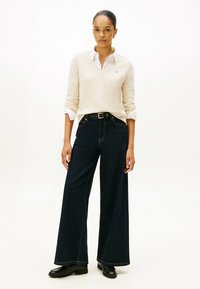 Vrouw gekleed in een beige V-hals trui over een wit shirt, met donkere wijde spijkerbroek, zwarte riem en zwarte loafers, staande tegen een effen achtergrond.