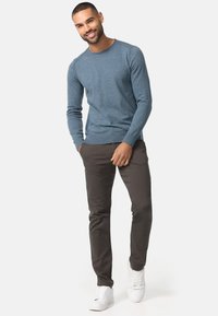 Pull en tricot bleu à manches longues, associé à un pantalon marron foncé et des baskets blanches. Le modèle se tient debout, mettant en valeur une coupe décontractée.