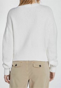 Suéter blanco de punto con dobladillo y puños acanalados, usado sobre pantalones beige claros con bolsillos traseros abotonados. La textura parece suave.