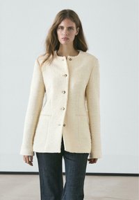 Cream-colored textured blazer with a round neckline, seven buttons, and side pockets. Worn over dark denim pants.