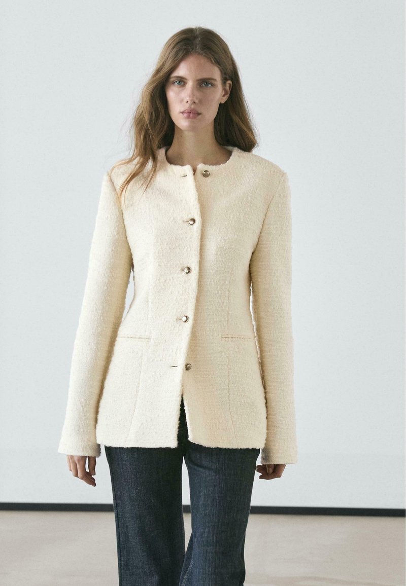 Cream-colored textured blazer with a round neckline, seven buttons, and side pockets. Worn over dark denim pants.