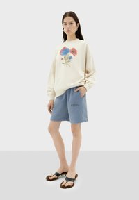 Crème sweatshirt met een bloemenverfontwerp, gecombineerd met blauwe shorts met een zak. Bruine fluffy flip-flops maken de outfit compleet.