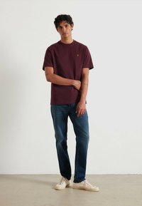 T-shirt girocollo bordeaux realizzata in tessuto morbido con maniche corte, abbinata a jeans blu scuro e sneakers di colore chiaro. Dettaglio del logo semplice sulla maglietta.