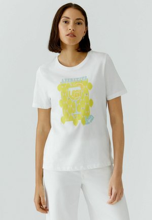 Frau trägt ein weißes T-Shirt mit gelbem Zitronenmuster und einer Cocktailglas-Grafik, kombiniert mit weißen Hosen, vor einem schlichten Hintergrund.