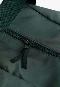 Dunkelgrüne Nylon-Tasche mit einem Reißverschlussfach. Der Reißverschluss hat passende grüne Ziehverschlüsse und einen strukturierten Tragegurt.