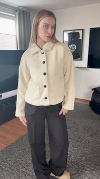 Γυναίκα σε εσωτερικό χώρο, ντυμένη με κρεμ φλις σακάκι με κουμπιά, μαύρο παντελόνι φαρδύ και μπεζ παπούτσια, στέκεται σε γκρι μοκέτα.