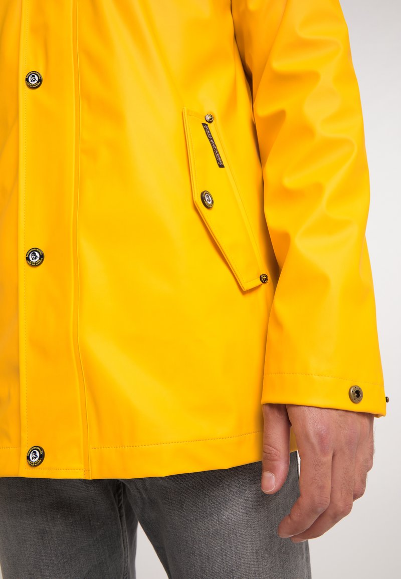 Gelbe wasserdichte Jacke mit Druckknöpfen und Taschenklappe, getragen über grauen Jeans, zeigt eine entspannte Hand an der Seite.