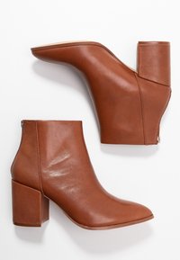 Bottines en cuir marron avec un bout pointu, un talon épais, et une fermeture éclair à l'arrière, présentant une texture lisse et brillante.
