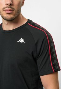 Melna T-krekls ar sarkaniem akcentiem uz piedurknēm, kurā ir balts logotips uz krūtīm un gluda, viegla auduma tekstūra.