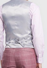 Gilet en satin argenté avec une texture lisse, présentant un détail de nœud dans le dos et une boucle ajustable, associé à une chemise à manches longues rose clair.